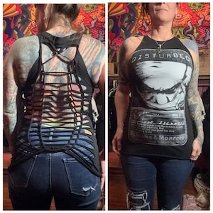 May include: Black tank top with a distressed design on the back. The front of the tank top features a graphic of a face with the word "DISTURBED" above it. The text "THE SICKNESS" is printed below the graphic.