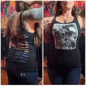 May include: Black tank top with a graphic print of a band and the word "RADIOHEAD". The back of the tank top has a unique cut-out design with crisscross straps. The person is wearing blue jeans.