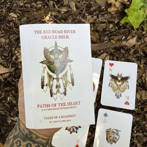 May include: A white oracle deck titled "The Red Road River Oracle Deck" with the text "Paths of the Heart" and "Tales of a Roadman." The deck includes a 54-card deck with a booklet. Several cards are visible, featuring illustrations and card suit symbols.