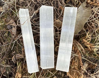 May include: Three translucent, rectangular selenite crystal wands are arranged on a bed of dried moss and twigs. The wands are vertically oriented, with a slightly textured surface, and appear to be glowing with an inner light. The natural setting enhances the crystals' ethereal quality.