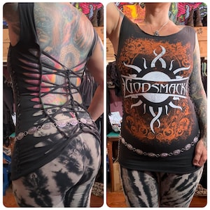 May include: A black sleeveless tank top featuring a large white and orange Godsmack graphic on the front. The back of the garment is heavily modified with a series of vertical cutouts and a crisscross lace-up design that runs down the entire length.