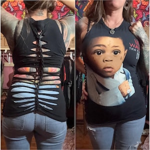May include: Black tank top with cut-out design on the back. The front of the tank top features a graphic of a baby. The model is wearing light blue jeans.