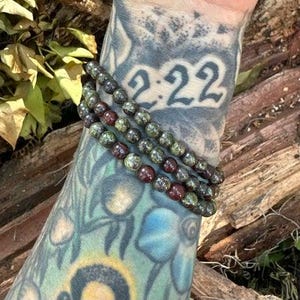May include: Three layered bracelets made of green and red gemstone beads. The bracelets are worn on a tattooed arm.