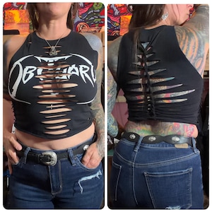 May include: Black, distressed tank top with the band name "Obituary" in white lettering. The front of the shirt has horizontal cut-outs. The back of the shirt also has cut-out detailing. The model is wearing blue jeans and a black belt.