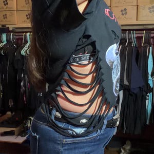 May include: Black distressed crop top with cut-out details. The shirt is paired with blue jeans and a studded belt. The image shows a glimpse of a tattoo on the arm. The background includes clothing racks and boxes.