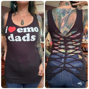 May include: A black sleeveless tank top featuring the white text "i" followed by a red heart symbol and the white text "emo dads" on the front. The back of the garment is designed with a series of horizontal cutouts that are woven together.