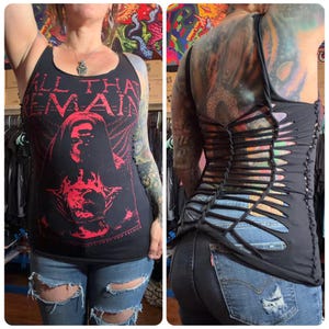 May include: Black tank top with red graphic text and image. The text reads "ALL THAT REMAINS" with an image of a person. The back of the tank top has a cut-out design with woven straps. The person is wearing ripped blue jeans.