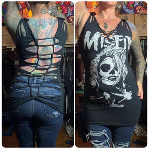 May include: Black tank top with a distressed design, featuring cut-out details and a laced neckline. The front displays a white graphic of a skull with the word "MISFIT" above it. The back of the top has a unique woven design.