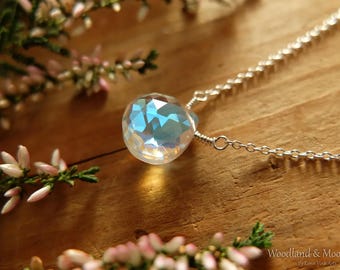 May include: A silver chain necklace with a clear iridescent gemstone pendant. The pendant is faceted and reflects light in a rainbow of colors. The necklace is photographed on a wooden surface with pink flowers.
