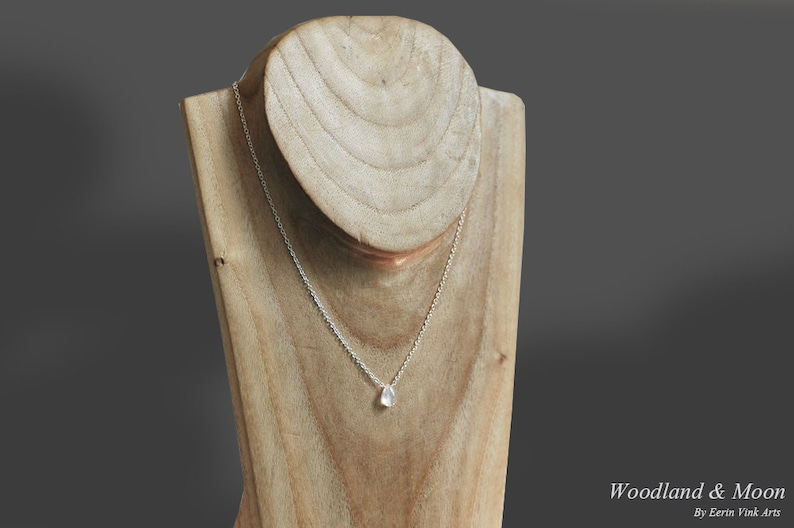 May include: A silver chain necklace with a teardrop-shaped pendant on a wooden display stand. The pendant is a milky white color.