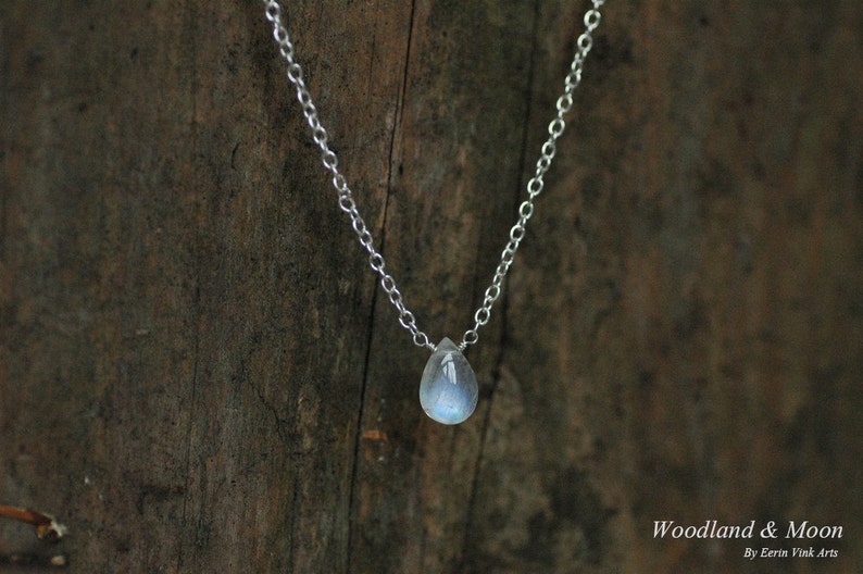 May include: A silver chain necklace with a teardrop-shaped moonstone pendant. The pendant is a light blue color with a shimmery finish. The necklace is hanging on a wooden surface.