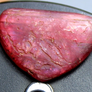 May include: A close-up of a pink and red gemstone with a rough, textured surface. The gemstone has a heart shape and is set against a dark gray background.