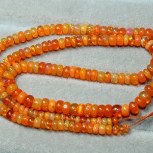 May include: A strand of round, orange fire opal beads arranged in a loose, multi-layered coil. The beads exhibit a translucent quality with subtle, shimmering internal flashes of green and yellow light, strung together on a thin orange thread.