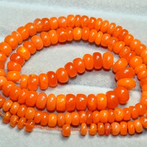 May include: A strand of vibrant orange, disc-shaped beads arranged in a loose, circular coil. The beads feature a smooth, polished finish with subtle variations in color intensity, strung together on a thin, matching orange thread.