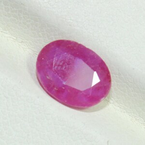 May include: A faceted, oval-shaped ruby gemstone. The gem is a vibrant pinkish-red color, with multiple facets that catch the light. The stone is set against a white background.