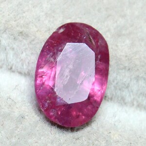 May include: A faceted, oval-shaped ruby gemstone. The gem features a deep pink hue with a lighter, almost colorless, central facet. The stone is set against a neutral, textured background, highlighting its color and cut.