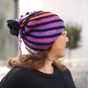 May include: A woman wearing a purple, black, and orange striped knit hat with a black bow on the side. The hat has a long tie that hangs down the back of her neck.