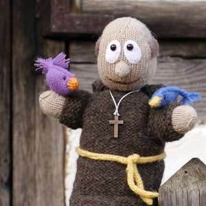 Può includere: Una bambola di lana vestita da monaco con una tunica marrone, una cintura gialla e una collana con una croce d'argento. La bambola tiene due piccoli uccelli di lana, uno viola e uno blu.