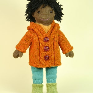 May include: A knitted doll with brown skin, black curly hair, and a smiling face. The doll is wearing a bright orange hooded sweater with brown buttons, teal blue pants, and green knitted boots.