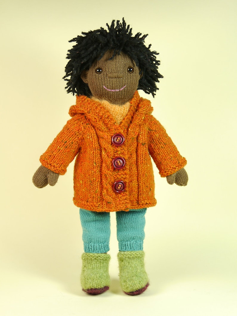 May include: A knitted doll with brown skin, black curly hair, and a smiling face. The doll is wearing a bright orange hooded sweater with brown buttons, teal blue pants, and green knitted boots.