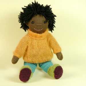 May include: A knitted doll with brown skin, black curly hair, a yellow sweater, and blue pants. The doll is sitting with its legs crossed and has a happy expression.