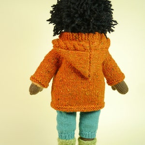 May include: A knitted doll with black curly hair wearing an orange hooded sweater and blue pants.