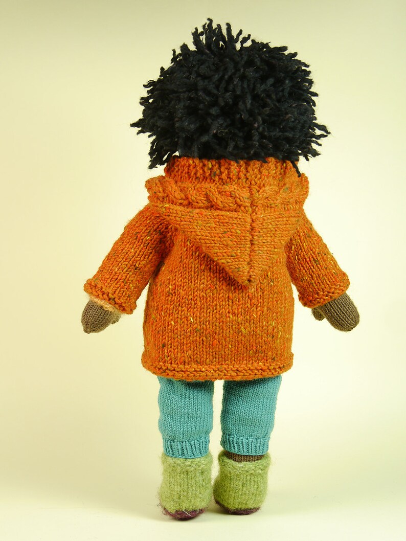May include: A knitted doll with black curly hair wearing an orange hooded sweater and blue pants.