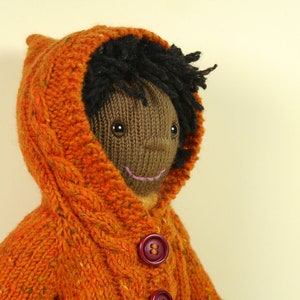 May include: A knitted doll with brown skin, black hair, and a smiling face. The doll is wearing a bright orange hooded sweater with a cable knit pattern and brown buttons.