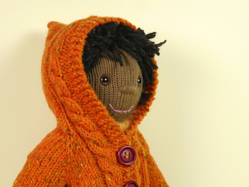 May include: A knitted doll with brown skin, black hair, and a smiling face. The doll is wearing a bright orange hooded sweater with a cable knit pattern and brown buttons.