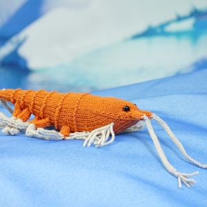 Könnte beinhalten: Ein gestricktes orangefarbenes Garnelenspielzeug mit weißen Antennen. Das Spielzeug liegt auf einer blauen Oberfläche.