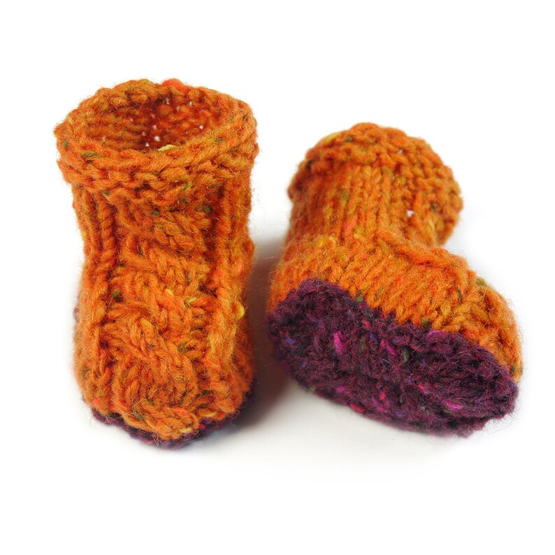 Puede incluir: Un par de botitas de beb&eacute; de punto naranja con ribete morado. Las botitas est&aacute;n hechas con un hilo grueso y tienen un patr&oacute;n de punto texturizado.