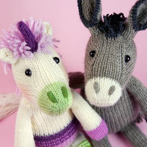 May include: A white knitted unicorn with a purple mane and a green and purple striped tail, next to a grey knitted donkey with black hooves.