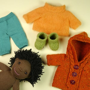 May include: A knitted doll with brown skin and black curly hair wearing a teal knitted pants, an orange knitted sweater, and a brown knitted jacket with a hood. The doll is also wearing green knitted booties.
