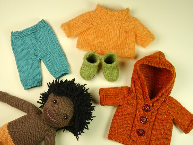 May include: A knitted doll with brown skin and black curly hair wearing a teal knitted pants, an orange knitted sweater, and a brown knitted jacket with a hood. The doll is also wearing green knitted booties.