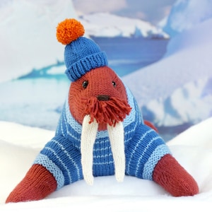 May include: A knitted walrus wearing a blue and white striped sweater and a blue knit hat with an orange pom-pom. The walrus is lying on a white surface.