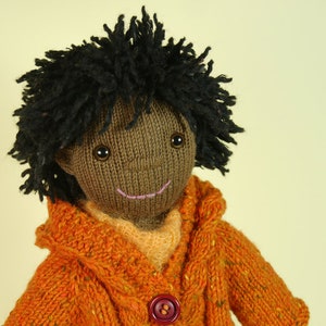 May include: A knitted doll with brown skin, black yarn hair, and a smiling face. The doll is wearing a bright orange knitted sweater with a button.