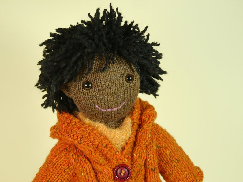 May include: A knitted doll with brown skin, black yarn hair, and a smiling face. The doll is wearing a bright orange knitted sweater with a button.