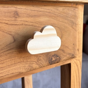 Könnte beinhalten: Ein heller Holz-Schubladenknopf in Form einer stilisierten Wolke mit sichtbarer horizontaler Holzmaserung. Der Knopf ist an der Vorderseite einer mittelbraunen Holzschublade mit ausgeprägter natürlicher Holzmaserung befestigt.