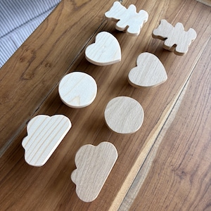 May include: A collection of eight light-colored wooden knobs in various shapes, including two bear silhouettes, two hearts, two circles, and two clouds. Each knob features a smooth, natural wood grain finish and a simple, flat-faced design.
