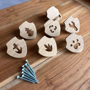 May include: A set of seven light-coloured wooden cabinet knobs, each shaped like a shield with a unique cutout design including an anchor, tree, recycle symbol, wrench, paw print, and leaf, accompanied by a small pile of silver metal screws.