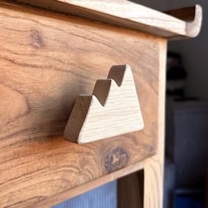 Puede incluir: Mueble de madera con un detalle en forma de montaña. El mueble está hecho de madera marrón claro, mostrando un patrón de vetas natural. El acento de la montaña es de un tono de madera más claro, creando contraste.