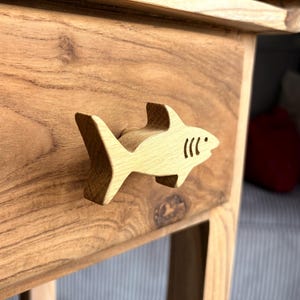 Könnte beinhalten: Ein hölzerner, haiförmiger Schubladengriff ist an einer Holzschublade befestigt. Der Hai hat eine Flosse, einen Schwanz und drei Kiemen. Das Holz hat eine natürliche Maserung und eine hellbraune Farbe. Die Schublade ist Teil eines Holztisches.