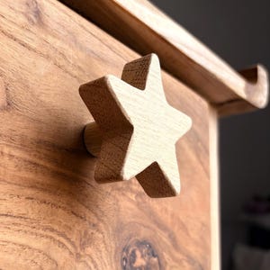 May include: Close-up of a wooden star-shaped drawer pull. The drawer pull is light brown and attached to a wooden drawer. The wood grain is visible on both the drawer and the star.