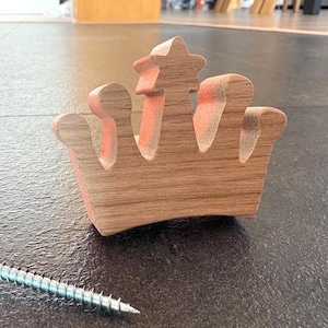 Puede incluir: Una corona de madera con una estrella en la parte superior, pintada con detalles en naranja. La corona es de madera marrón claro y tiene un diseño clásico. Un tornillo plateado está en primer plano.