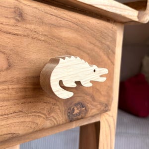 Op de afbeelding: Een houten kast met een lichtgekleurde, alligatorvormige handgreep. De kast is gemaakt van warm getint hout en de alligatorhandgreep heeft een klein, zwart stipje als oog. Het ontwerp van de kast is eenvoudig en natuurlijk.
