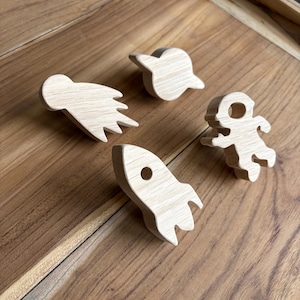 May include: Four wooden space-themed toys: a rocket, a spaceship, an astronaut, and a satellite. The toys are made of light-colored wood and are arranged on a wooden surface.