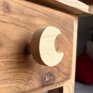Könnte beinhalten: Nahaufnahme einer Holzschublade mit einem halbmondförmigen Griff. Schublade und Griff sind aus hellem Holz gefertigt, das die natürliche Maserung zeigt. Der Griff ist mit einem kleinen, runden Stift an der Schublade befestigt.