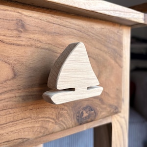 Op de afbeelding: Houten lade met een handvat in de vorm van een zeilboot. De lade en het handvat zijn gemaakt van lichtbruin hout. De zeilboot heeft een eenvoudig ontwerp met een driehoekig zeil en een horizontale basis. De lade maakt deel uit van een houten meubelstuk.