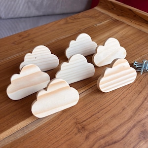 Peut inclure: Un ensemble de sept boutons en bois clair non fini en forme de nuage avec des motifs de grain de bois visibles. Chaque nuage présente un design arrondi à plusieurs lobes. Un petit tas de vis en métal argenté est placé à droite des pièces en bois.