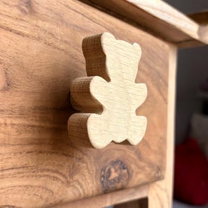 Peut inclure: Tiroir en bois avec un bouton en forme d'ours en peluche sculpté. Le tiroir est en bois brun clair avec un grain visible. Le bouton en forme d'ours est également en bois et fixé à la façade du tiroir.